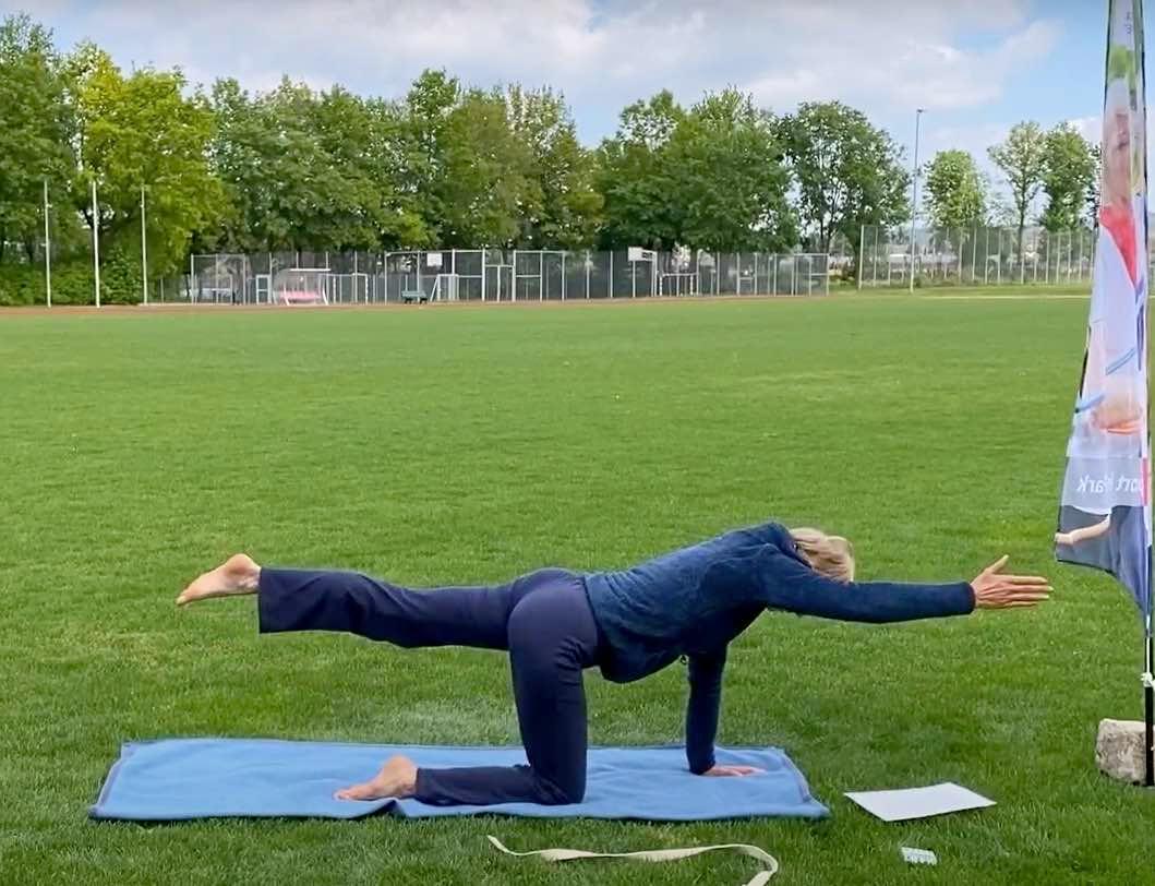 Eine Dame, die im Rahmen von "Sport im Park" eine Pilates-Übung zeigt. Sie streckt auf einer Yoga-Matte ein Bein und eine Hand aus und hält dabei ihren Rücken gerade parallel zum Boden.