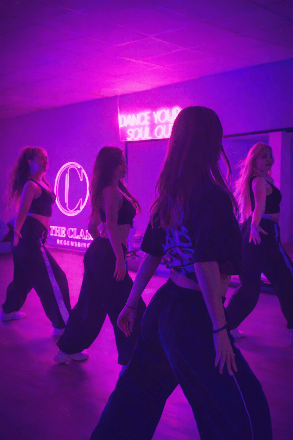 Vier junge Frauen tanzen in dem Tanzstudio vor dem Spiegel. An der Wand hängen Neonschilder mit dem Schriftzug "Dance your soul out" und "THE CLASS Regensburg".